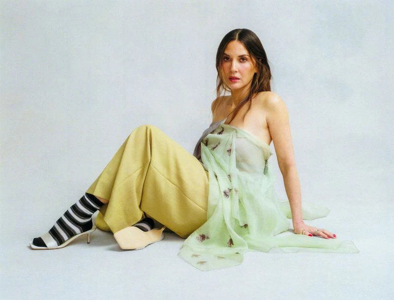 A woman with long dark hair, wearing a green shawl and yellow pants, sits on the floor with striped socks and heeled shoes in a studio setting.