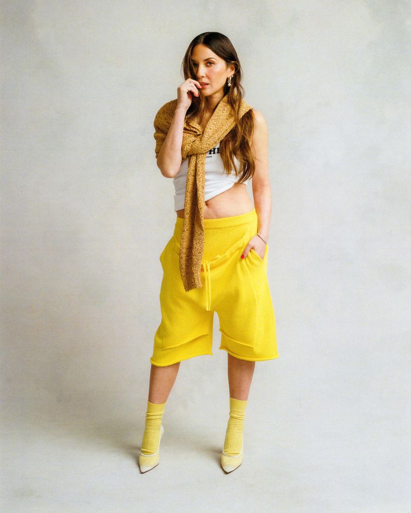 A woman stands in a photo studio wearing bright yellow culottes, a white top, yellow shoes, and a beige shawl, set against a neutral backdrop.