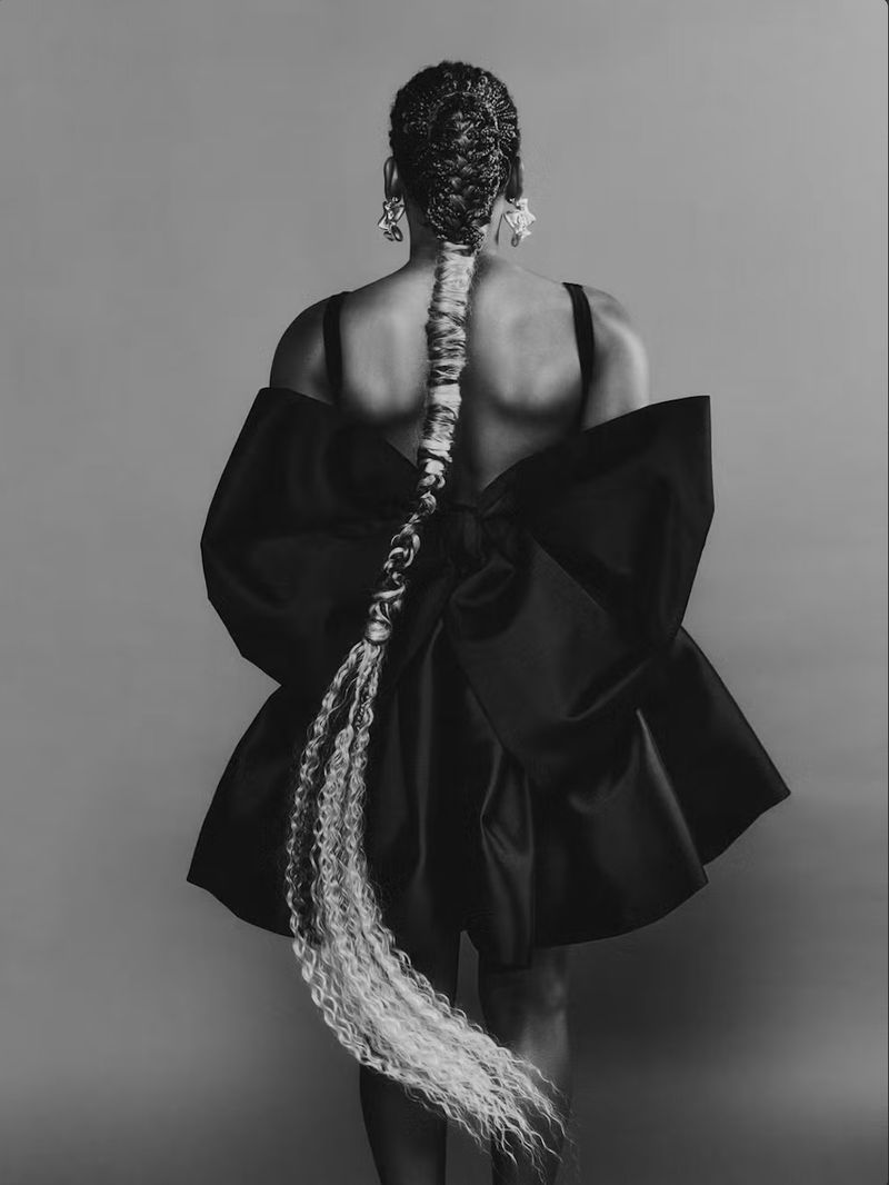 Back view of an adult female with a long braided hairstyle, statement earrings, and a black dress with an oversized bow detail.