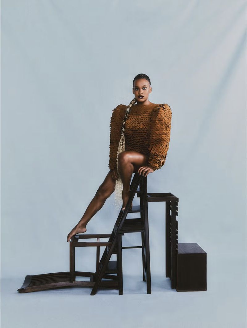 Woman with braided hair and patterned dress sits atop a dark wooden staircase in a photo studio against a gradient background.