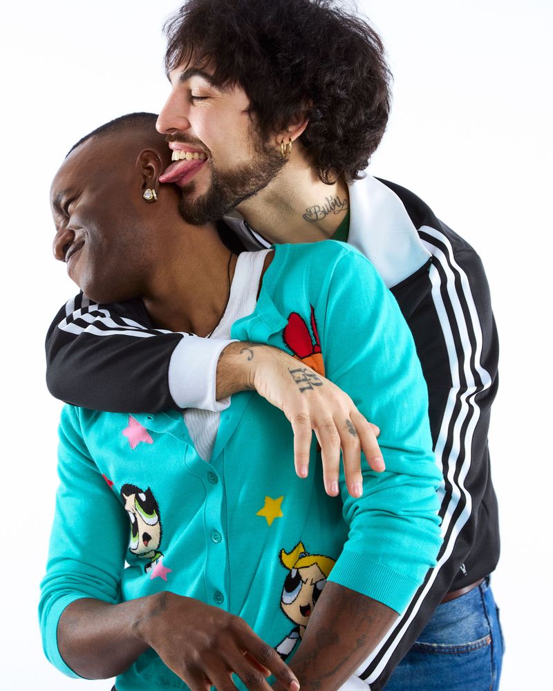 Two men embrace indoors; one man licks the other's ear while wearing a light blue Powerpuff Girls cardigan and jeans against a white backdrop.