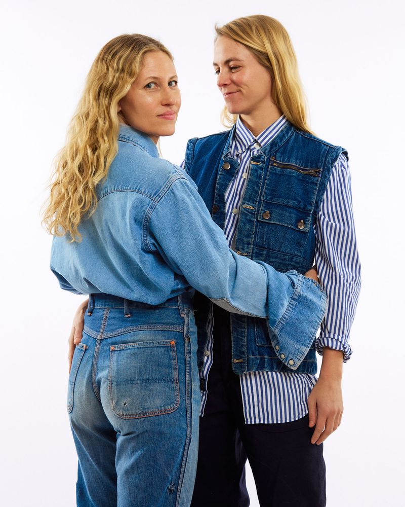 Two adult women in denim and striped shirts pose in a studio setting, one looking at the camera, the other gazing at her companion.
