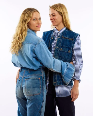 Two adult women in denim and striped shirts pose in a studio setting, one looking at the camera, the other gazing at her companion.