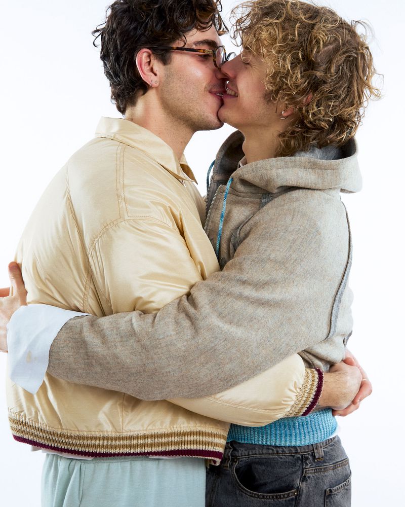 Close-up of two adult men embracing and kissing indoors against a white background, wearing casual clothing such as jacket and hooded sweatshirt.