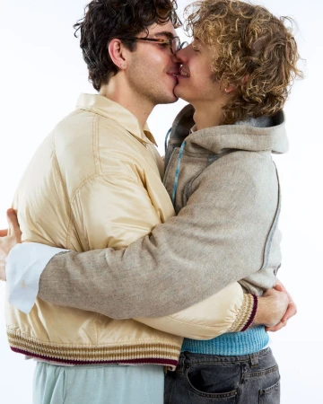 Close-up of two adult men embracing and kissing indoors against a white background, wearing casual clothing such as jacket and hooded sweatshirt.