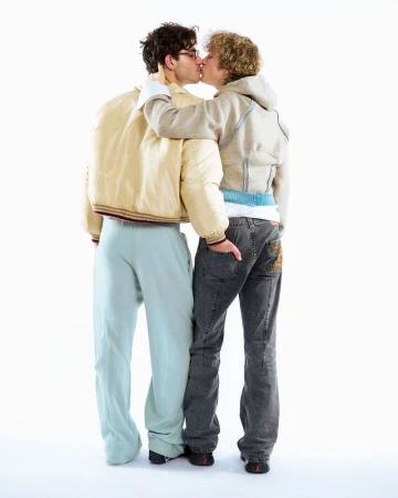 Two men, viewed from behind, embrace and kiss while standing in a white studio. One wears jeans and a hoodie, the other sweatpants and jacket.