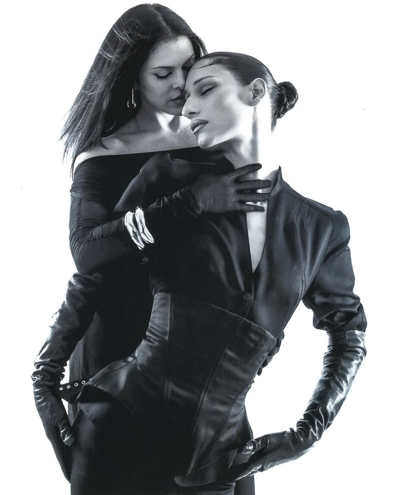 Two women in elegant black fashion, one with her hands on the other's neck, in a monochrome studio portrait against a white backdrop.