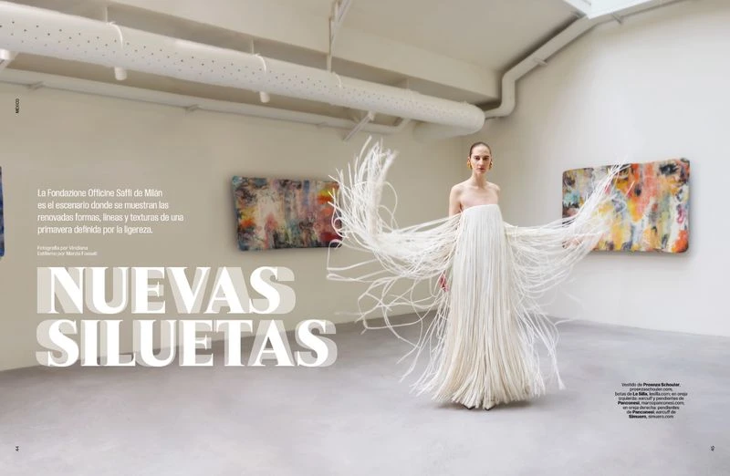 Fashion editorial shot of a model in a floor-length white fringe dress, posing in a gallery with modern art on the walls, shot indoors.