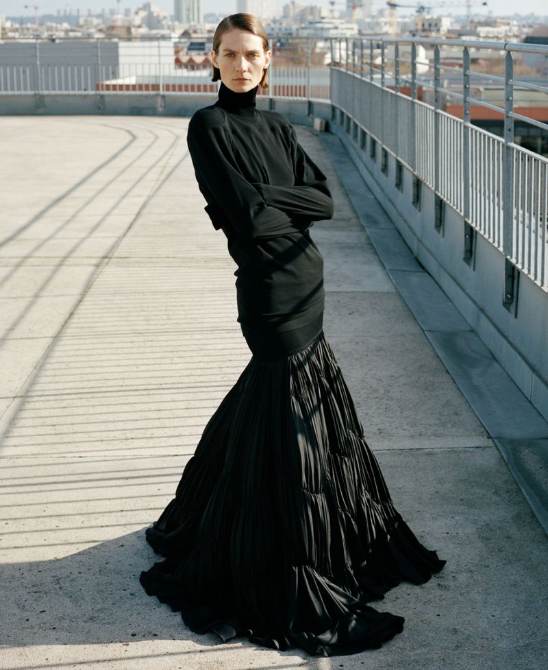 A slender woman poses in a long black dress on a rooftop with cityscape views, showcasing a high-fashion look.