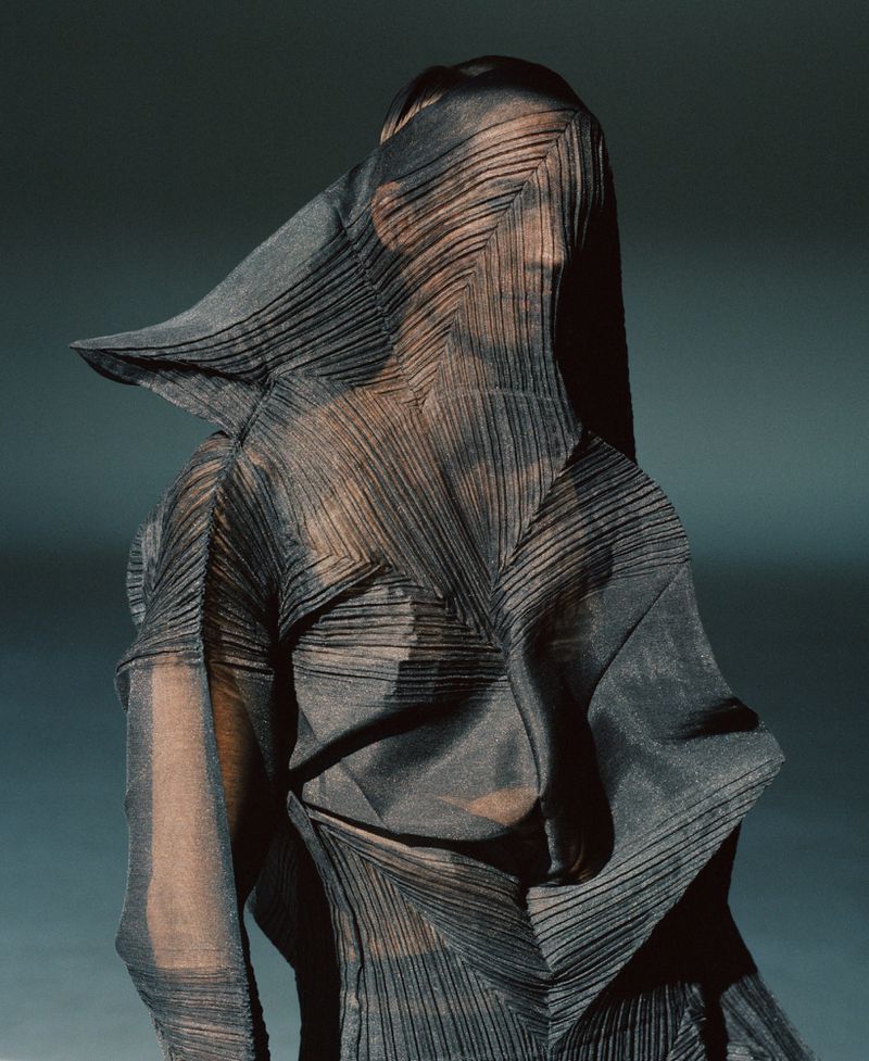 Fashion shot featuring a person draped in a geometrically pleated garment, face obscured, set against a dark grey-blue backdrop in a studio.