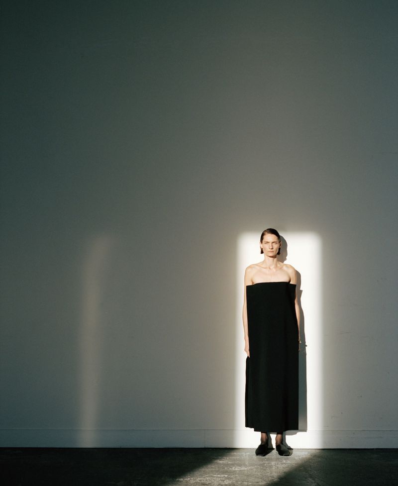 Fashion photography of a woman in a black dress standing against a neutral wall with light and shadow creating a studio-like atmosphere.