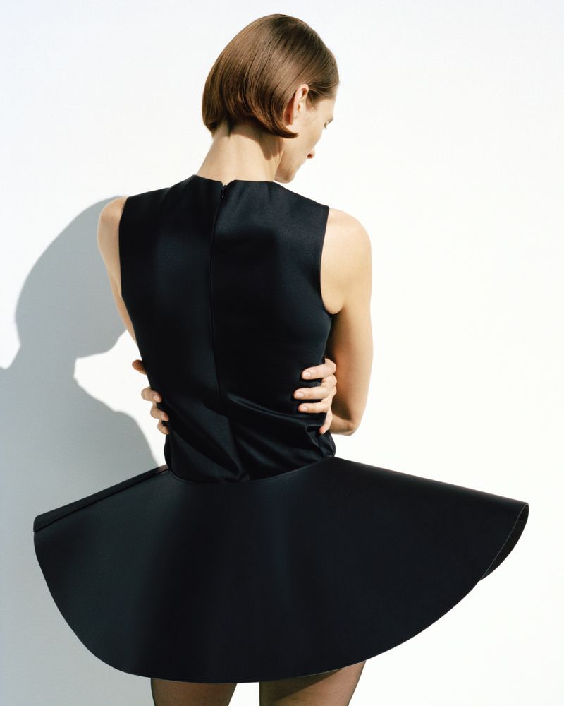 Back view of a woman with short brown hair in a black, sleeveless dress with a circular skirt standing in a brightly lit studio.