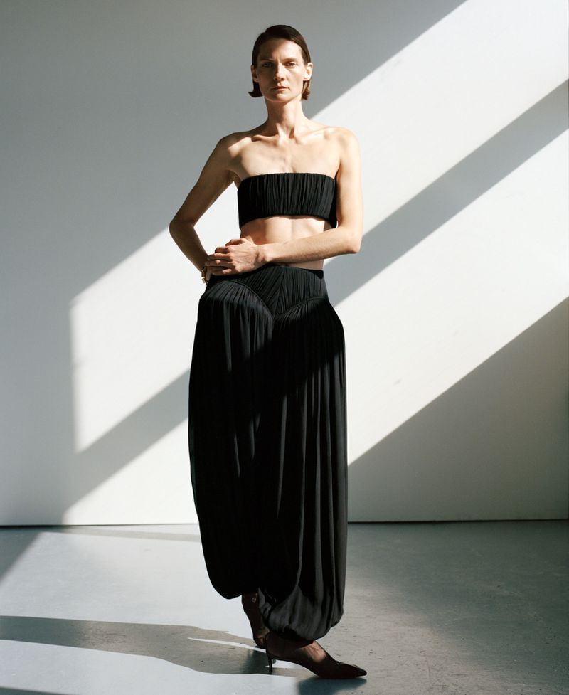 A slim woman poses in a strapless black crop top and voluminous black pants in a studio with natural light creating shadows.