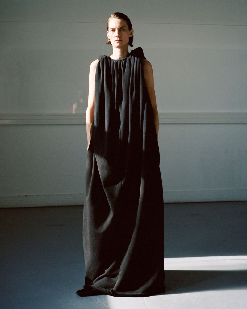 A female fashion model poses in a long black gown with a bow detail, standing in a photo studio with natural light. 