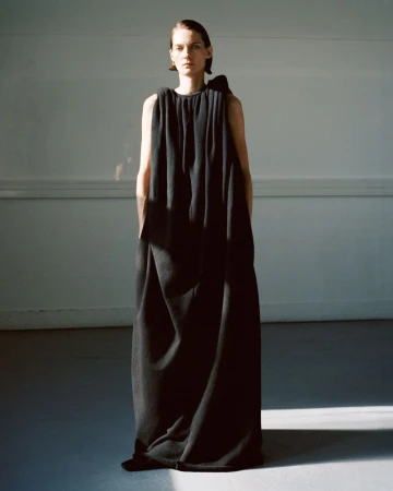 A female fashion model poses in a long black gown with a bow detail, standing in a photo studio with natural light. 