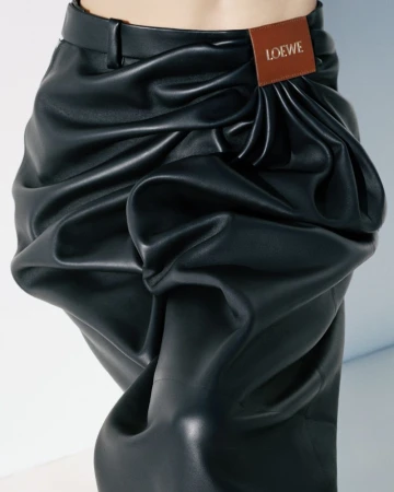 Close-up of a person wearing black faux leather pants with draped detailing and a brown leather LOEWE brand patch at the waist.