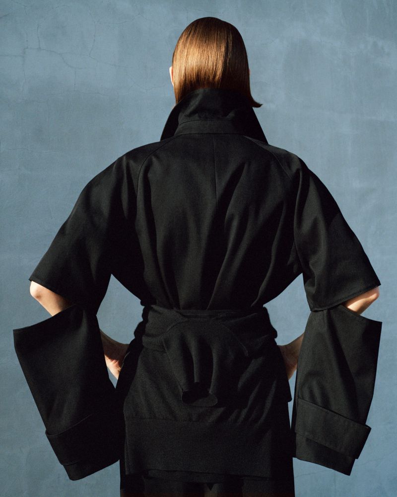 Back view of a woman with short brown hair wearing a black jacket-dress with a belt against a textured blue background.