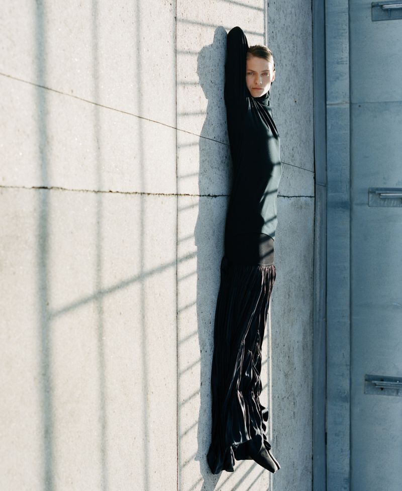 Fashion shot: Woman in long black dress poses against a concrete wall with dramatic shadows creating a high-fashion editorial image.