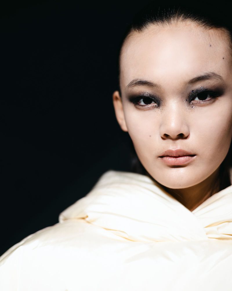 Close-up portrait of an East Asian woman with smoky eyeshadow wearing a puffy, quilted cream-colored jacket against a dark background.