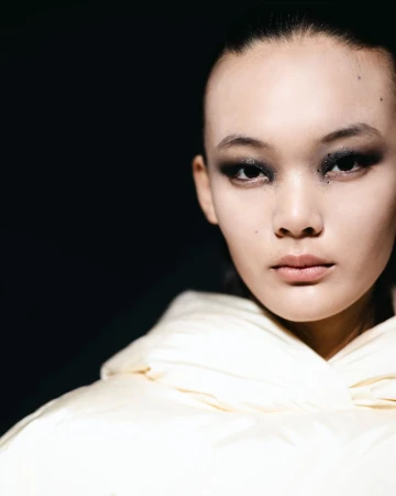 Close-up portrait of an East Asian woman with smoky eyeshadow wearing a puffy, quilted cream-colored jacket against a dark background.