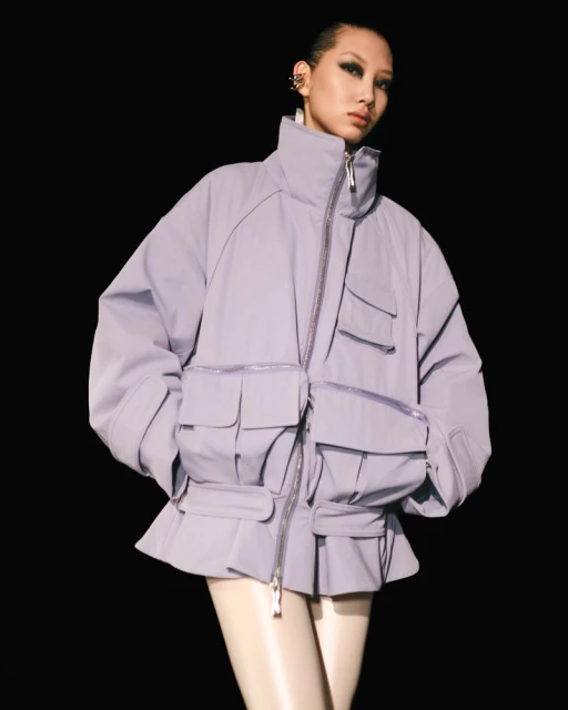 Fashion shot of an Asian female model posing in a lavender puffy coat with large pockets against a black backdrop.