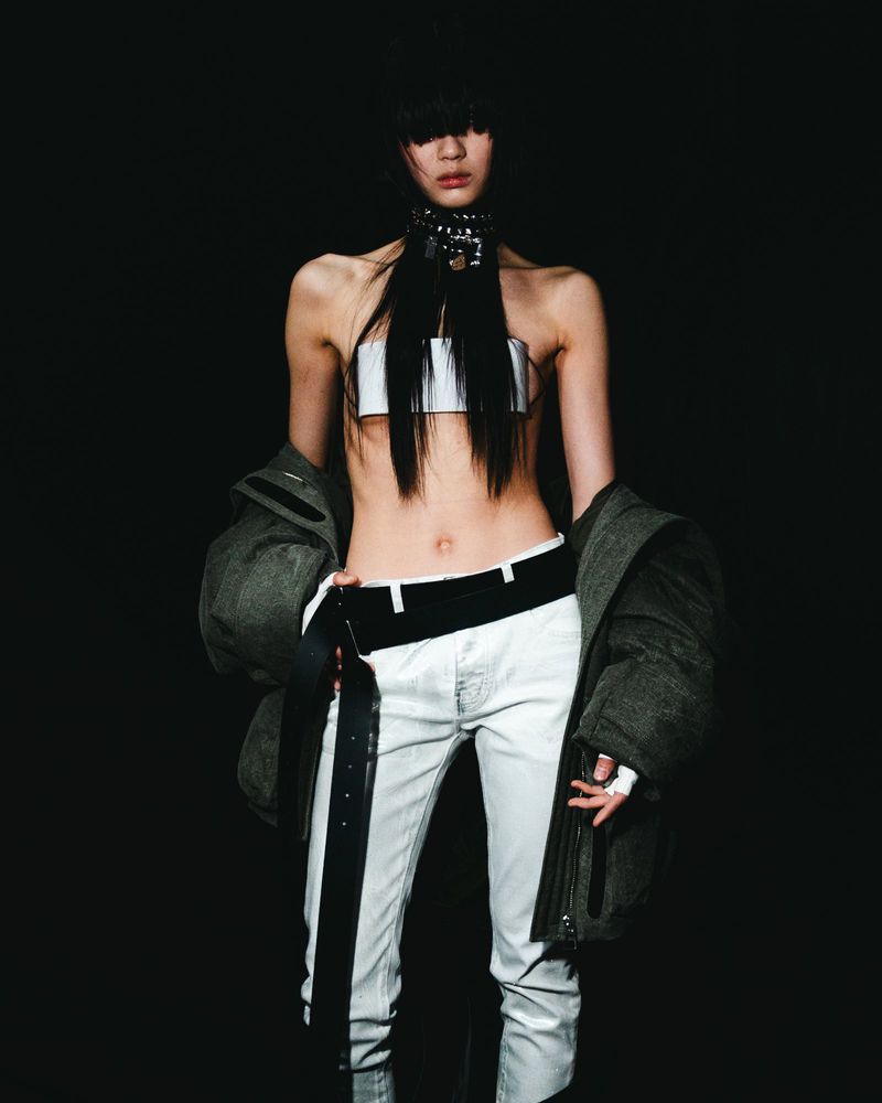 Fashionable adult female poses in a photo studio wearing a white top, pants, and a bulky jacket against a dark background.