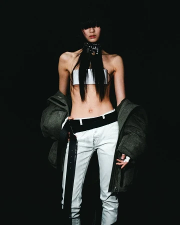Fashionable adult female poses in a photo studio wearing a white top, pants, and a bulky jacket against a dark background.