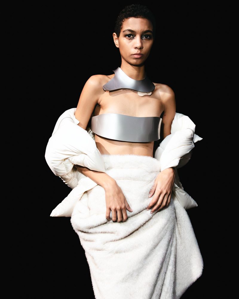 Model showcases futuristic silver metal top and collar paired with a white down jacket and a furry towel on a black studio backdrop.