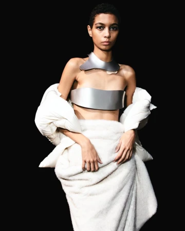Model showcases futuristic silver metal top and collar paired with a white down jacket and a furry towel on a black studio backdrop.