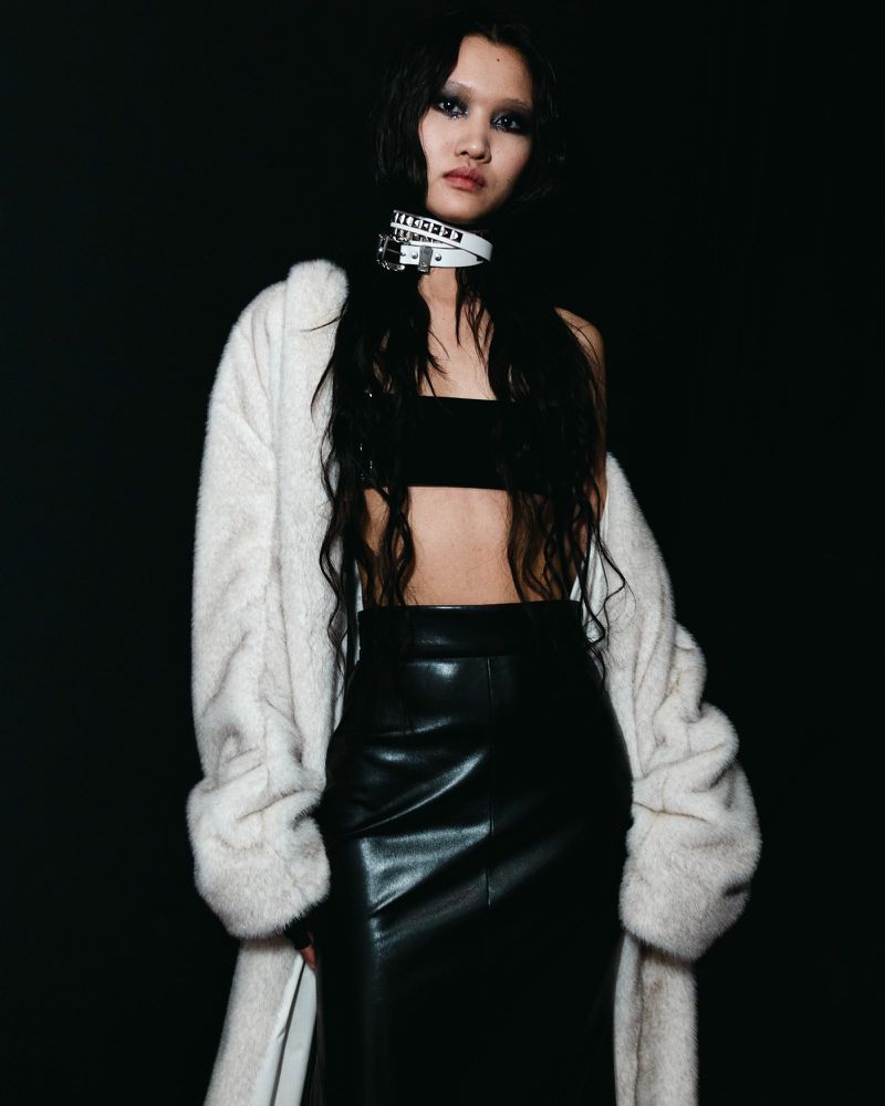 A slender woman with dark hair poses in a black top and shiny black pencil skirt, wearing a fur coat and choker in a studio setting.