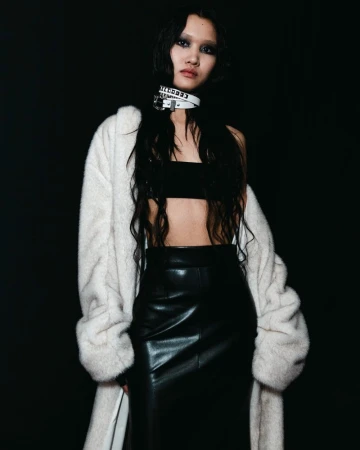A slender woman with dark hair poses in a black top and shiny black pencil skirt, wearing a fur coat and choker in a studio setting.