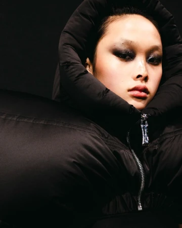 Portrait of Asian woman with dark eye makeup wearing a large black puffy jacket with a silver zipper against a dark backdrop.