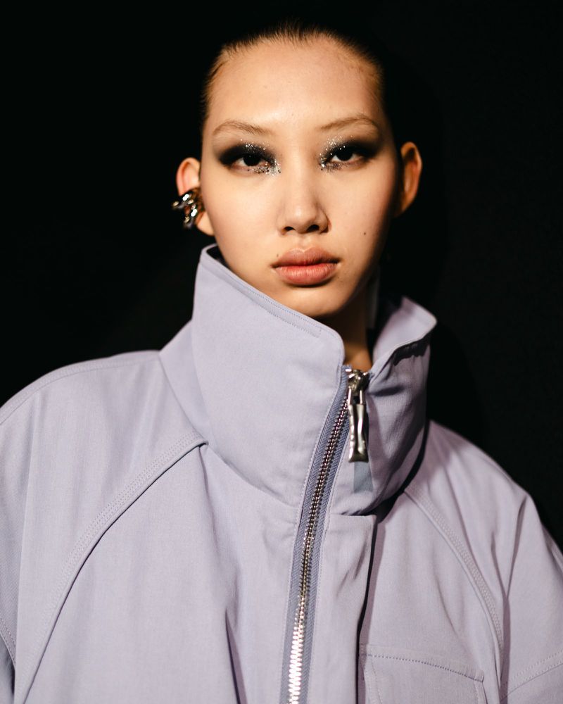 Close-up shot of a woman with unique eye makeup, wearing a light purple high-collared jacket, and a silver ear cuff, against a dark background.