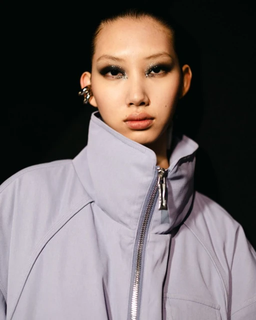 Close-up shot of a woman with unique eye makeup, wearing a light purple high-collared jacket, and a silver ear cuff, against a dark background.