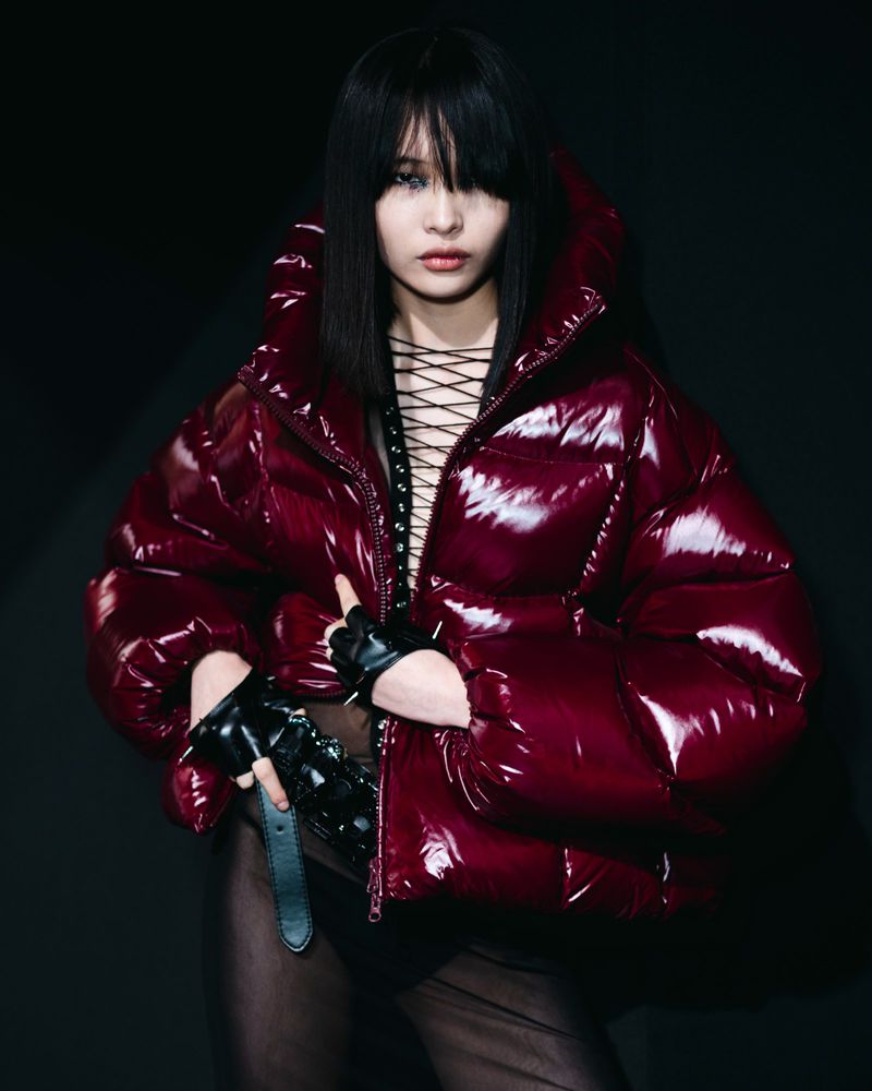 Young woman with bangs poses in a glossy red puffer jacket, black top and gloves against a dark background in a fashion-style portrait.