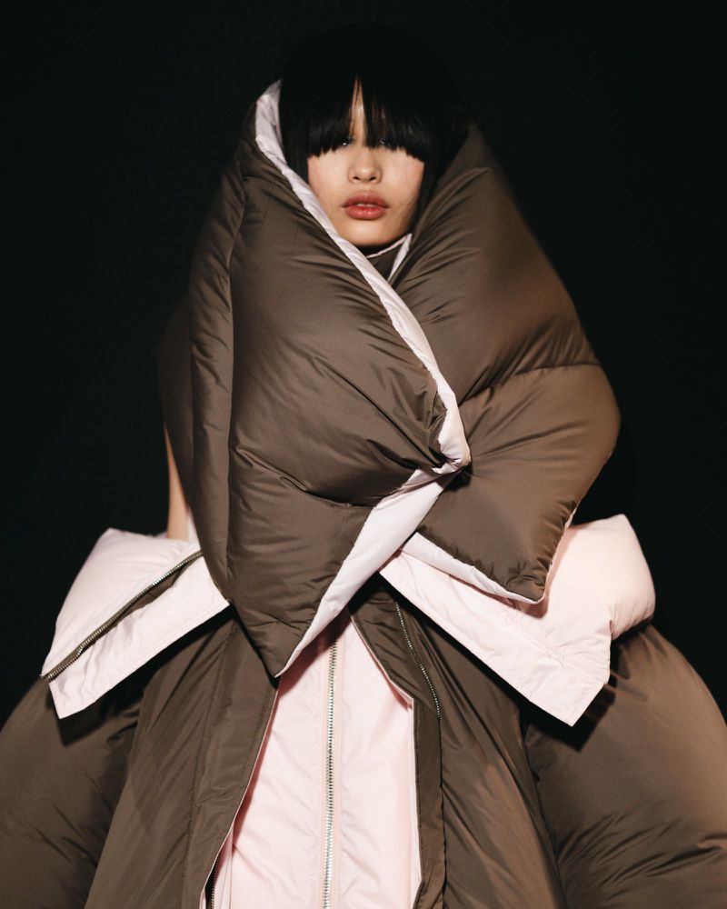 Woman in an oversized brown down puffer coat, posing against a dark backdrop, showcasing a unique fashion statement with visible fringe.