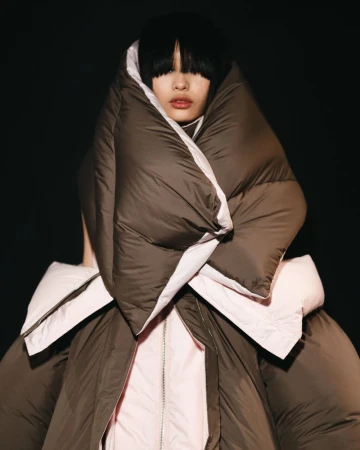 Woman in an oversized brown down puffer coat, posing against a dark backdrop, showcasing a unique fashion statement with visible fringe.