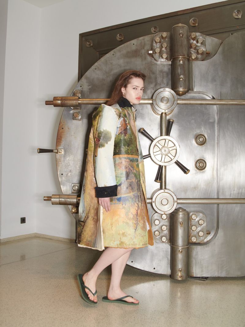 A woman stands in front of a large metal vault door wearing an earth-toned dress-coat and flip-flops, in an indoor, light-colored setting.