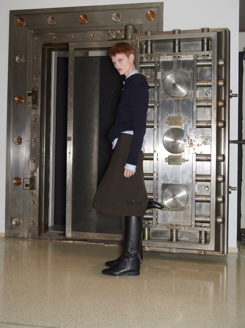 A slender adult stands in front of an open bank vault, wearing a navy sweater, brown skirt, and tall black boots in a full shot.