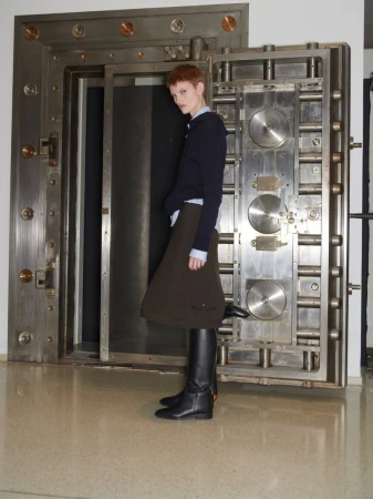 A slender adult stands in front of an open bank vault, wearing a navy sweater, brown skirt, and tall black boots in a full shot.