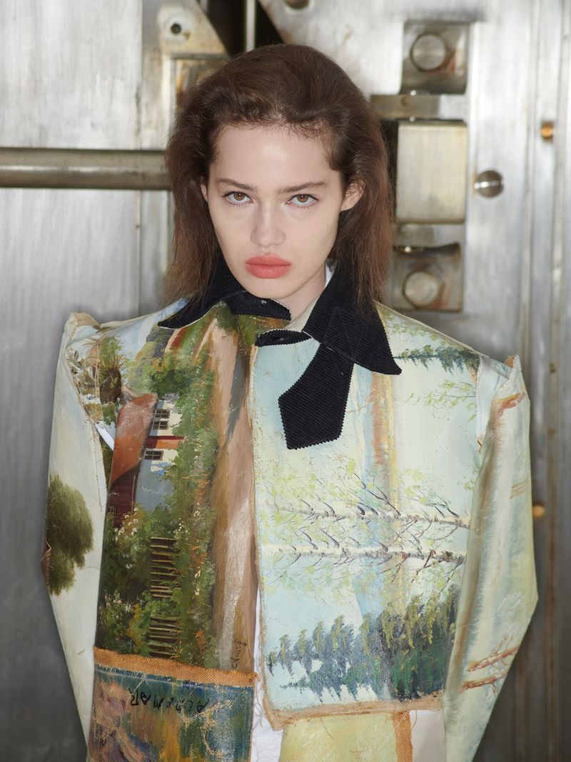A fashionable adult female in an artful painted jacket with a black collar poses indoors against a textured metal background.