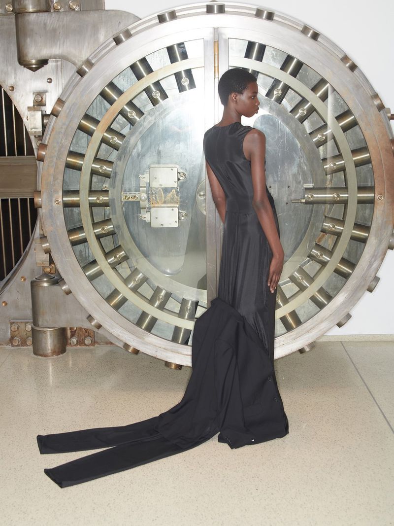An African American woman in a black dress with a long train stands in front of a large, round metal bank vault door in a studio setting.