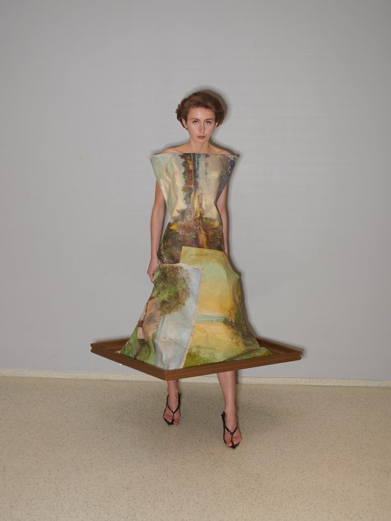 Woman in an art-inspired fashion dress with a landscape art print, framed by a square wooden frame at the hem, standing in a photo studio.