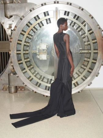 Model standing in front of a massive circular metal vault door wearing a dark sleeveless gown for Hodakova S/S 25