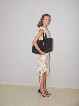 Model standing against a plain grey wall wearing a patterned cream dress and holding a large black woven handbag.
