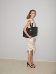 Model standing against a plain grey wall wearing a patterned cream dress and holding a large black woven handbag.