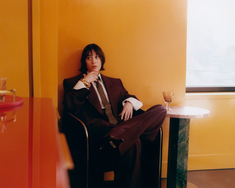 Adult woman in a maroon suit poses indoors with a glass of amber liquid on a table and an orange wall as a backdrop.