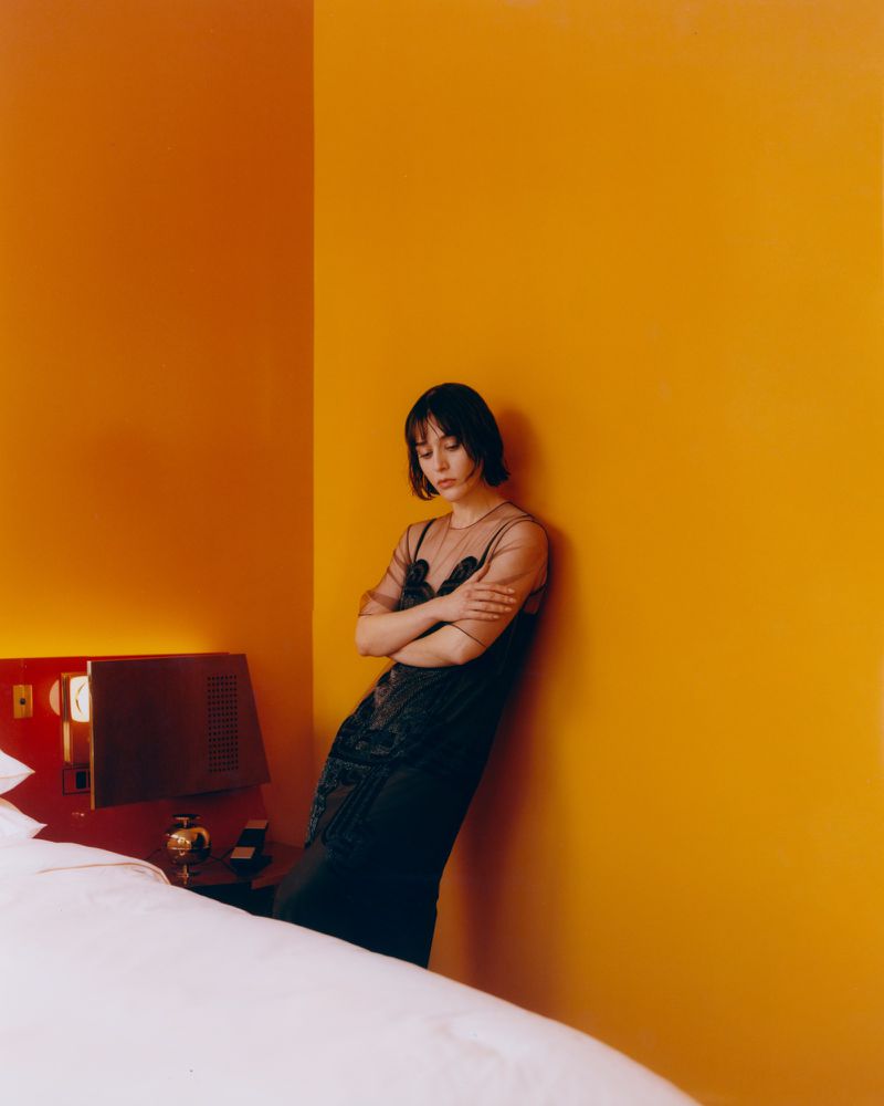 A slender woman with short dark hair leans against a bright orange wall in a minimalist hotel room setting, wearing an elegant black dress.