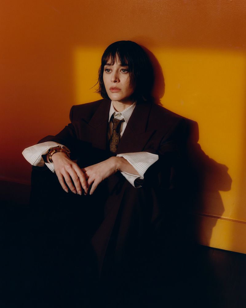 Elegant woman in an oversized suit and tie sits on a floor against a yellow wall in a studio. Portrait-style fashion photography.
