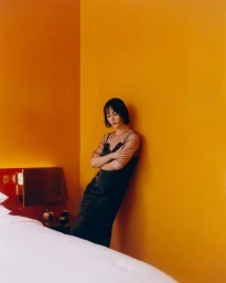 Lizzy Caplan leans against a vibrant yellow wall wearing a sheer black dress next to a white hotel bed.